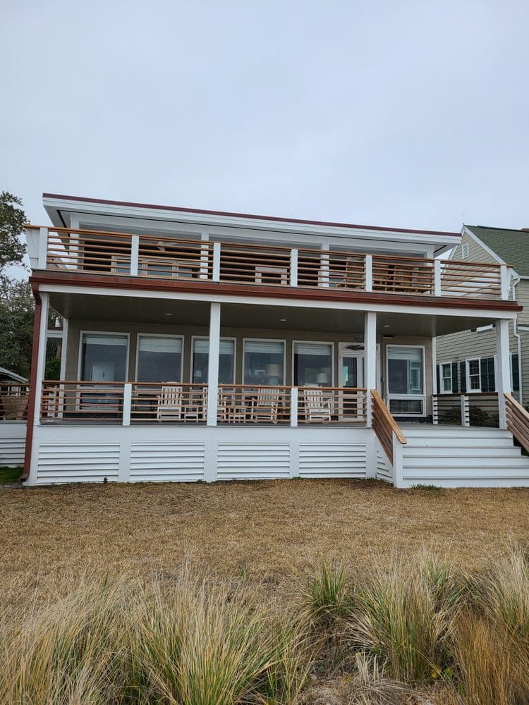 Modern coastal house with a spacious deck and rocking chairs, surrounded by natural landscaping.