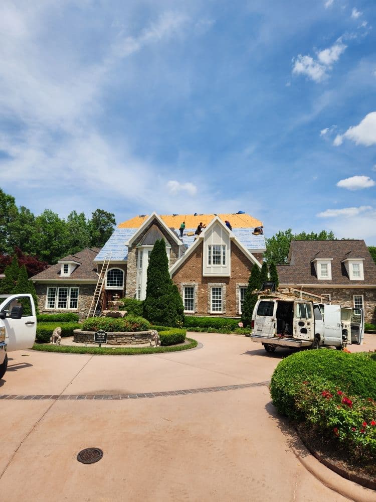 Roofing contractors working on a luxury home surrounded by manicured landscaping on a sunny day.