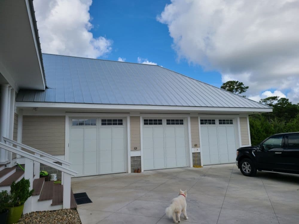 Modern garage with white doors, metal roof, and a dog in a sunny residential area.